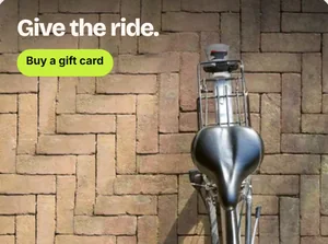 Een ontwerp voor een cadeaukaart: een fietszadel tegen een bakstenen muur onder de kop Give the ride, met een knop Buy a gift card.